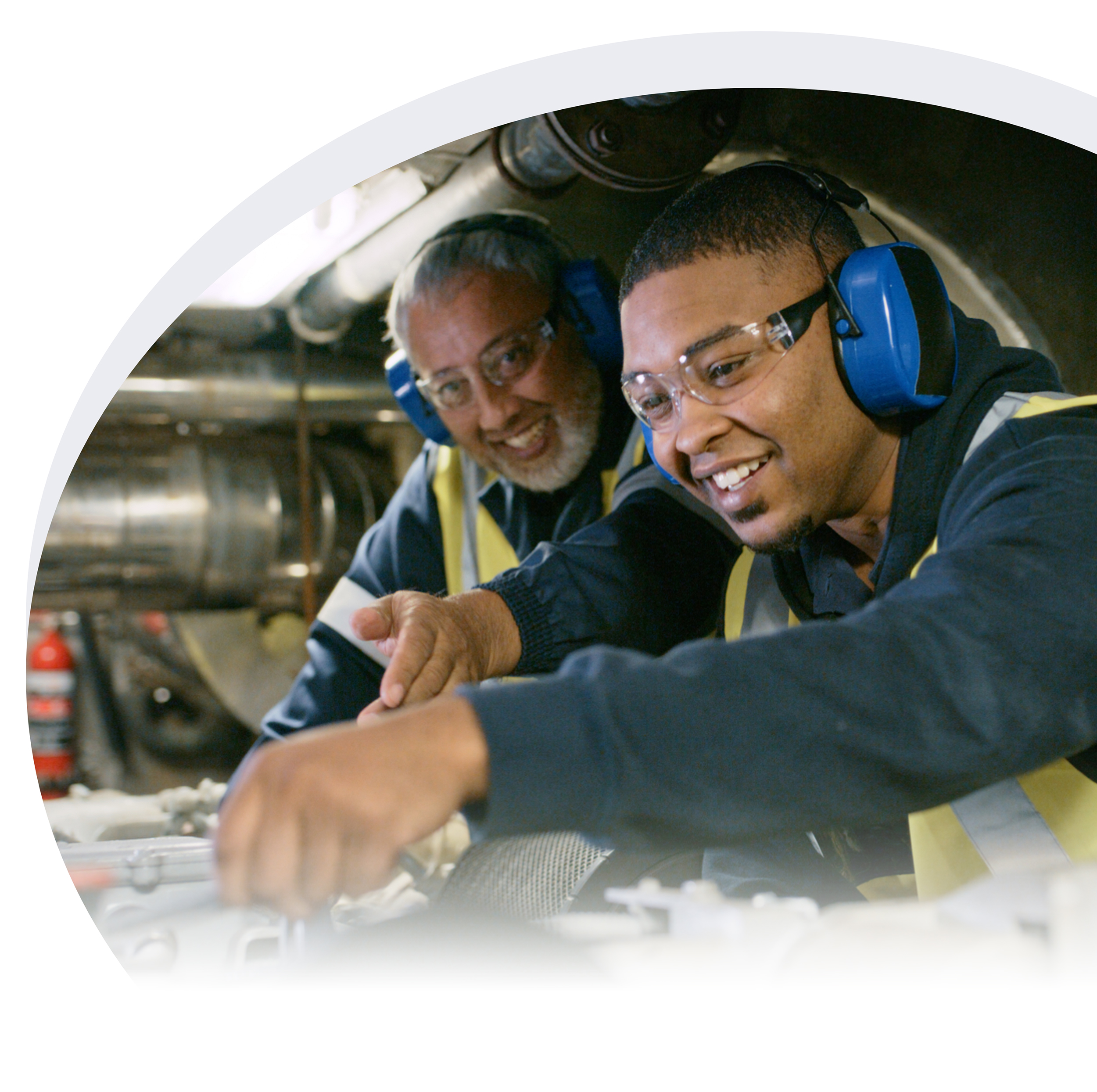 An older technician trains a younger technician. Both are wearing protective gear in a machine shop.