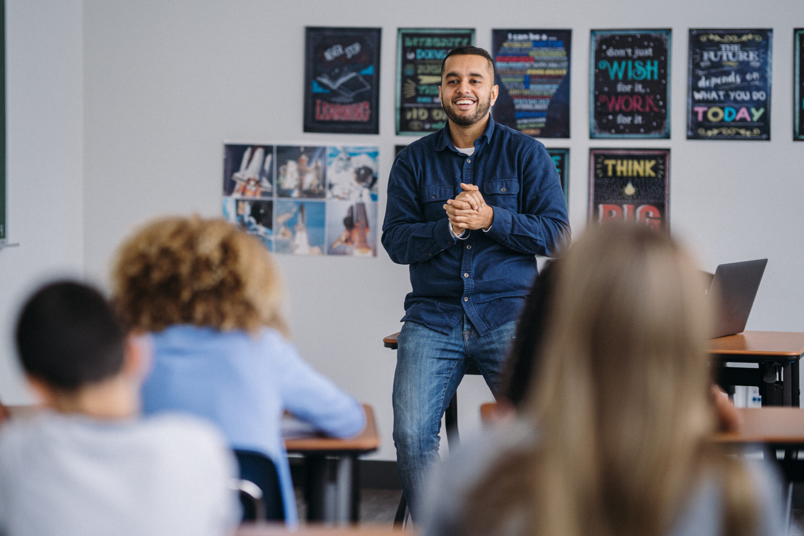 A male teacher of Hispanic ethnicity stands in the front of his middle school classroom, teaching a lesson to a group of concentrated students.