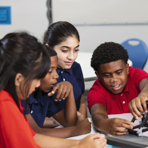 A multiracial group of middle school students in science class with their teacher examining a model of a bridge.